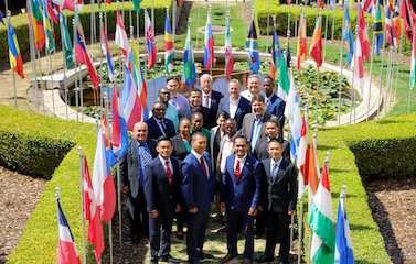 a picture of a group of participants in formal attires standing along side with each other in front of a beautiful green garden with various flags