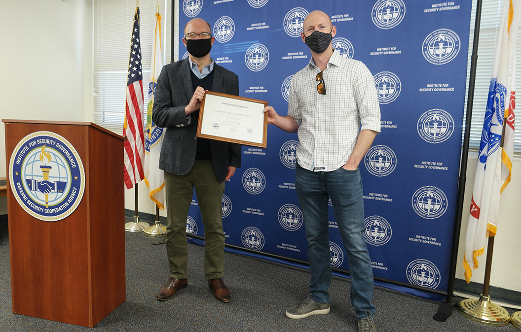Two men in facemasks standing in front of a blue banner, each holding one side of a certificate