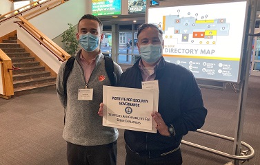 Two men stand shoulder to shoulder. They both wear face masks. The taller of the two wears a light grey zip up sweater. The other wears a navy blue wind jacket and holds a sign that says Institute for Security Governance, Strategies and Capabilities for Cyber Challenges.