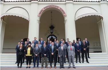 The class, their instructors, and the organizers of the event stand in three neat rows on the front steps of a building. The building has text above the steps in an arch that says, Naval Postgraduate School. The men are wearing suit coats. There are only two women standing in front. They are also dressed in formal attire.