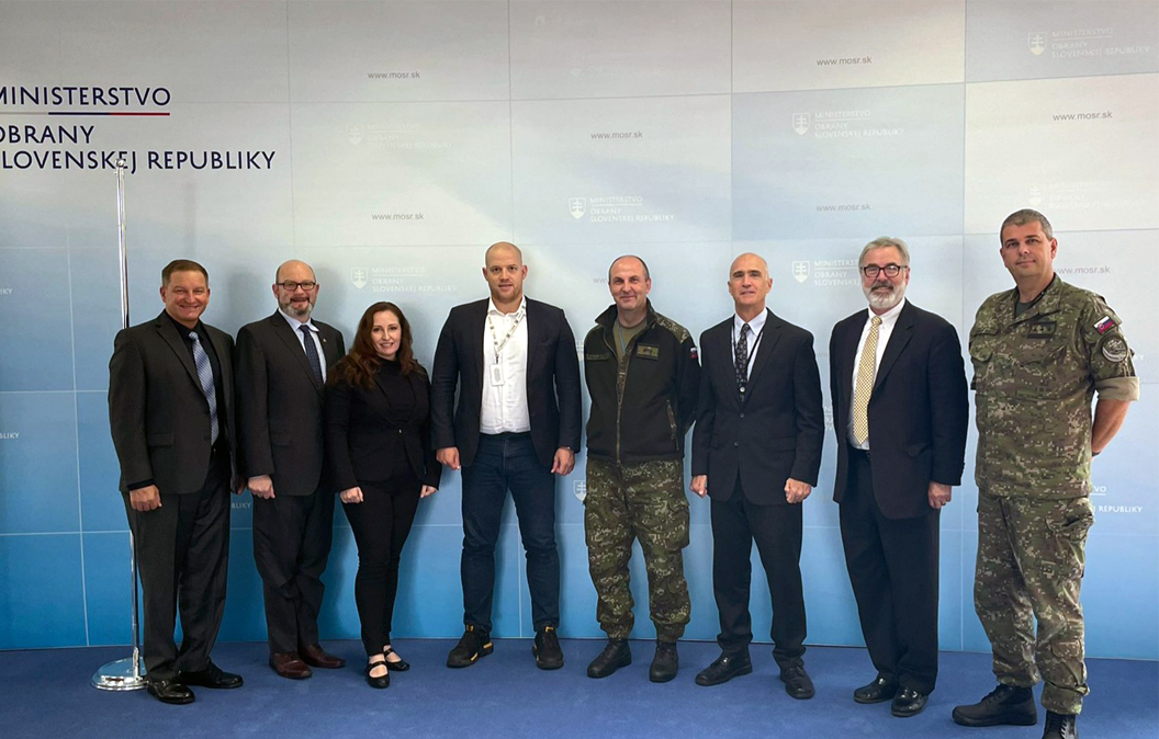 Seven men and one woman stand and pose in front of a step and repeat background with a Slovakian Embassy Logo and Title. The men and woman appear in various states of dress from suits to military fatigues.