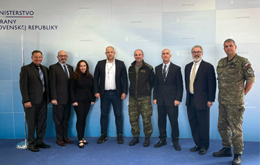 Seven men and one woman stand and pose in front of a step and repeat background with a Slovakian Embassy Logo and Title. The men and woman appear in various states of dress from suits to military fatigues.