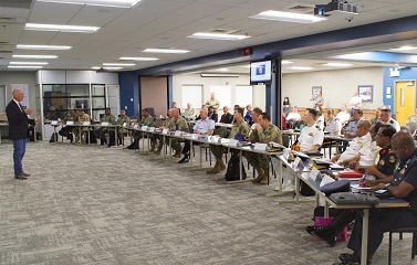 A man in a dark blue suit is speaking to a classroom of individuals. The class members each have tent cards in front of them. They sit at long tables arranged in a semicircle.
