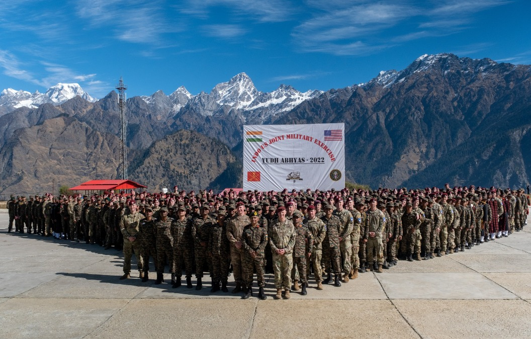 100+ people in military uniform stand in formation outside against the backdrop of a snowcapped mountain and blue skies. A sign behind them displays various country flags and text reading: UNDO Military Exercises Yudh Abhyas 2022