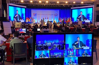 Looking out over a computer monitor showing the room being recorded. In a conference setting with flags and a speaker on the stage.