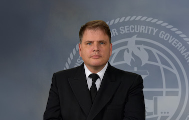 a picture of a man in a black suit sitting in front of a gradient grey ISG logo background