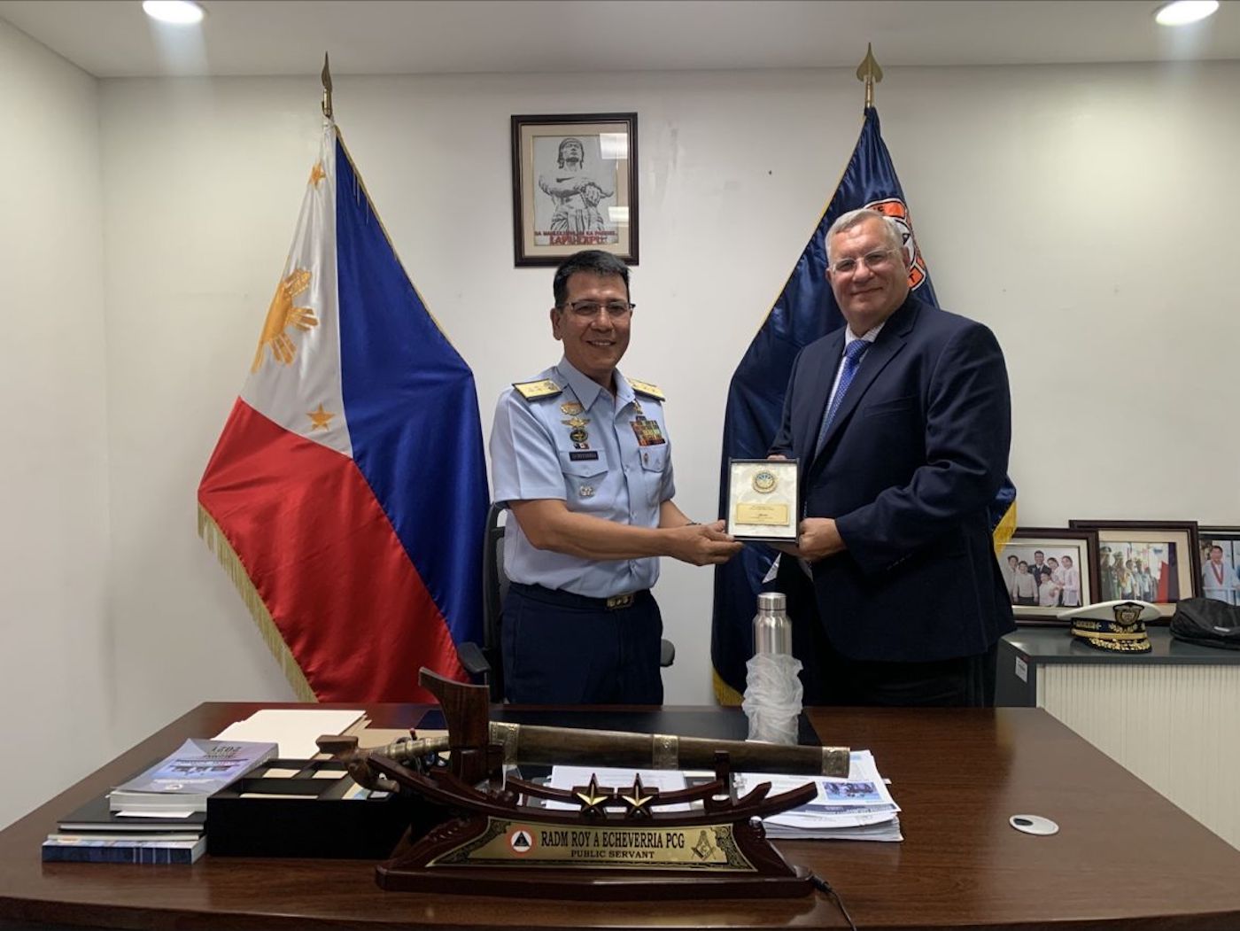 a picture of an officer handing an award to the man in a navy suit with the Philippines flag in the background