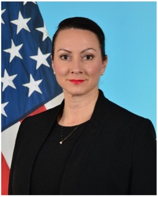 Woman in a suit, wearing a necklace, with her hair tied back standing in front of the U.S.A. flag