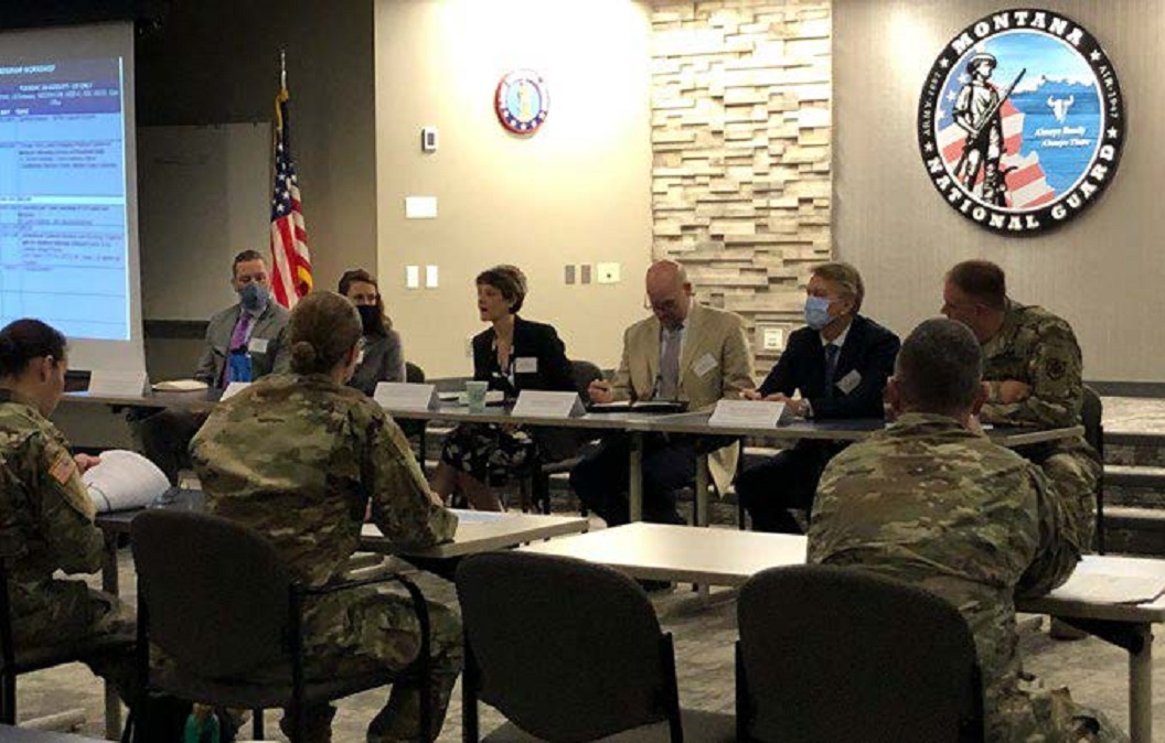 Several people sit facing each other in active discussion. Some are dressed in military fatigues while others are dressed in professional attire. Against the wall in the background is a large sign that says Montana National Guard.